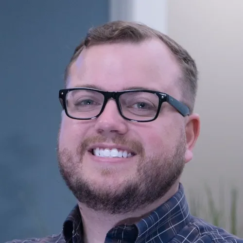 A man with short hair, a beard, and glasses smiles at the camera while wearing a checkered shirt.