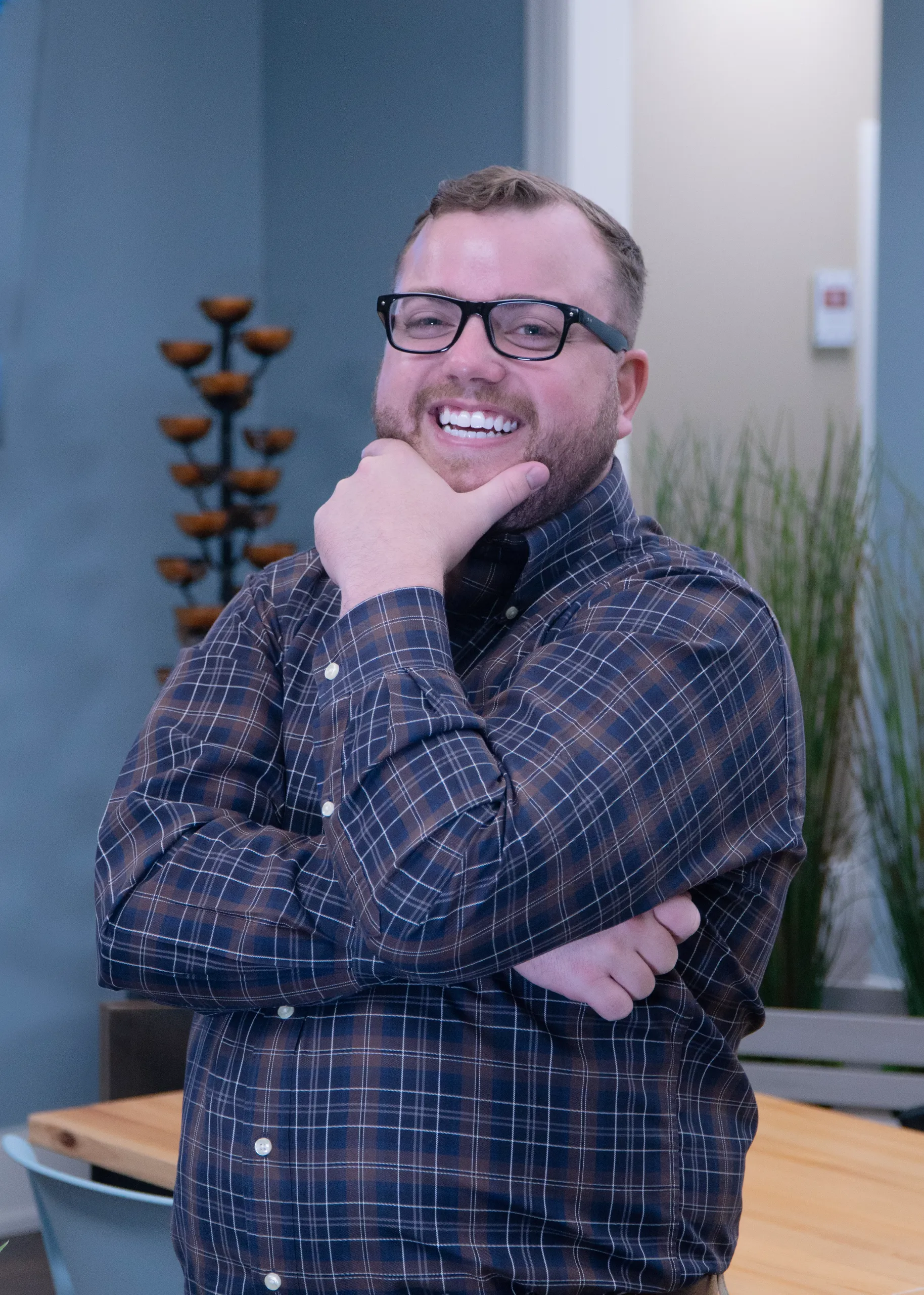 Man wearing glasses and a plaid shirt stands indoors, smiling with one hand on his chin and the other arm crossed, with plants and modern decor in the background.