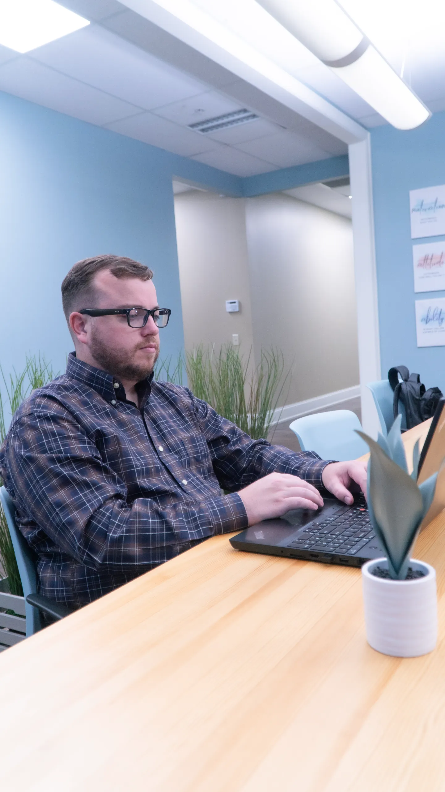 Man wearing glasses sits at a wooden table, typing on a laptop in a modern office with light blue walls and green plants nearby.