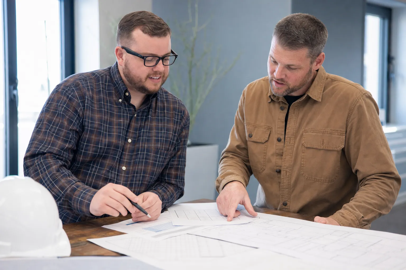 Two men discuss architectural blueprints at a table. One points at the plans while the other holds a pen. A white hard hat is on the corner of the table.