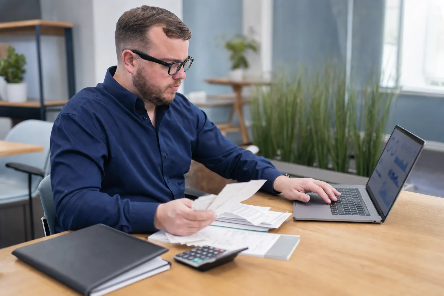 Man in blue shirt working at desk with a laptop, calculator, documents, and folder. He is holding papers and reviewing information on the computer.